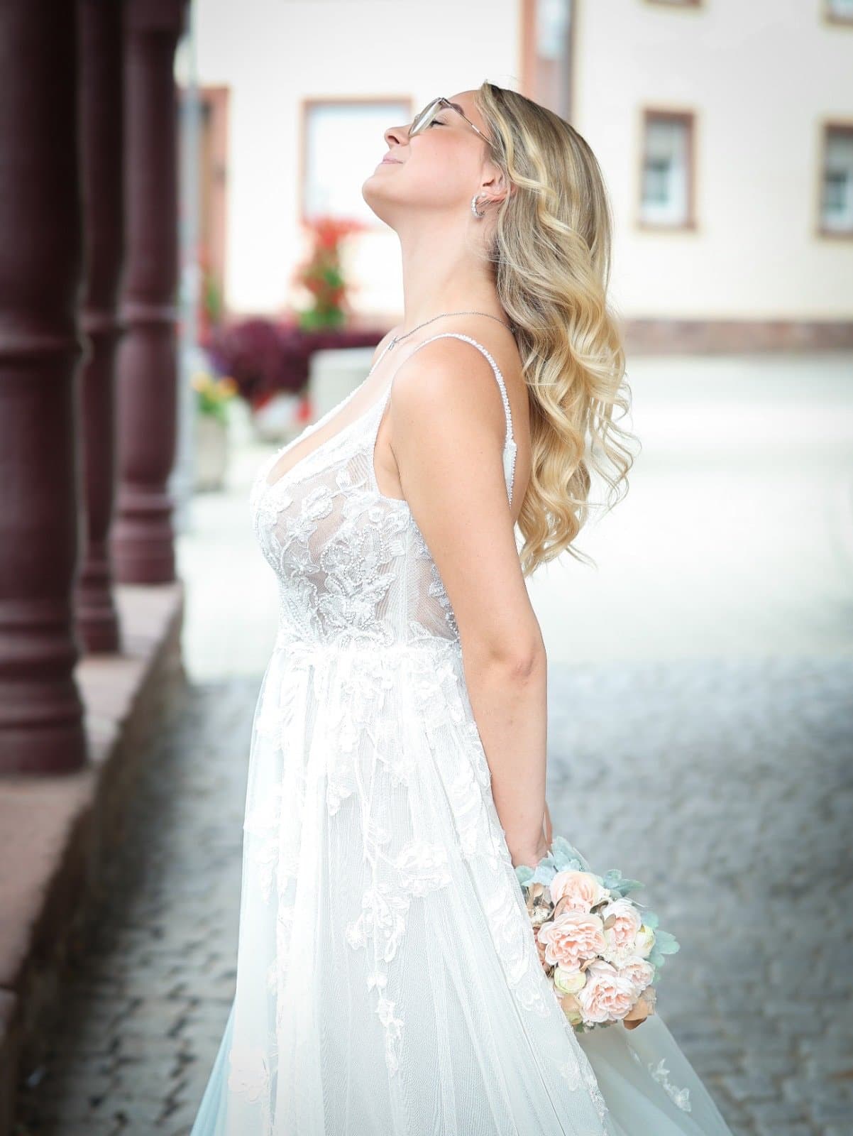 Lachende Braut mit Brautstrauß im Spitzenkleid unter Arkaden, Neue Kollektion von Brautmode Liebesgeschichte in Königsbach-Stein